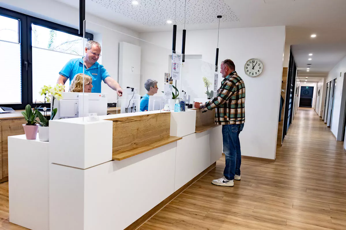 Gesprächssituation am Empfang: Arzt bespricht sich mit Mitarbeiterin im Vordergrund; Mitarbeiterin empfängt Patient im Hintergrund